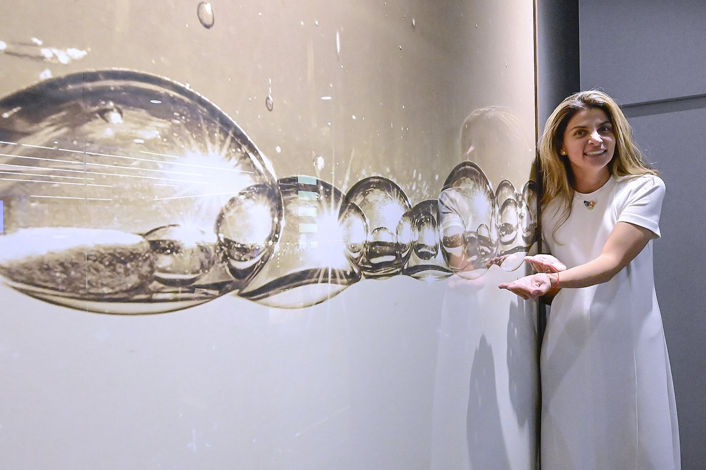 Mujer rodeada de copas de agua en una exposición, rodeada de un fondo oscuro y una pared gris